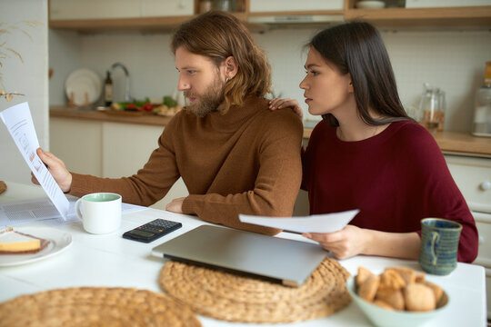Indoor shot of serious stylish couple of freelancers working together from home, sitting at dining table using laptop, calculator, studying documents. Young married family managing finances