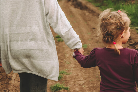 Mom And Daughter Are Holding Hands. View From The Back. The Sisters Walk Holding Hands. Portrait Of A Child In Nature. Copy Space. Beautiful Blonde Girl 4 Years Old.