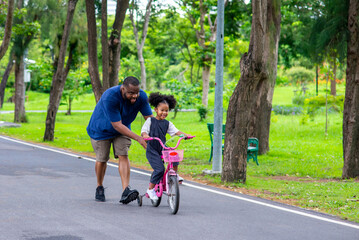 Happy mixed race family in park. African father teaching little daughter to ride a bicycle in the park. Dad and child girl kid having fun together outdoor lifestyle activity in weekend vacation.