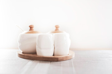 Fototapeta na wymiar White porcelain seasoning containers for salt, pepper and sugar. Wooden details. The set of kitchenware on a table. Selective focus on a wooden top, blurred background.
