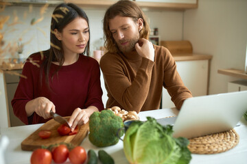 Cute young Caucasian couple cooking dinner following instructions in video recipe using laptop....