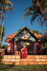 Portrait of a Buddhist monk sitting in front of a small temple.