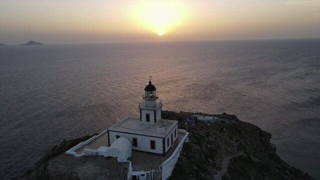 Aerial Panning Shot Of Akrotiri Lighthouse With Tourists Against Sky, Drone Flying Forward Over People On Rocky Mountain By Sea At Sunset - Santorini, Greece