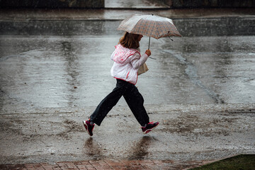 a girl with an umbrella runs through puddles in the rain. wet feet and clothes during a heavy...