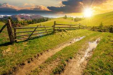 mountainous rural landscape at sunset in spring. path through grassy field. wooden fence on rolling hills. snow capped ridge in the distance. wonderful countryside scenery in evening light