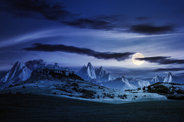 castle on the hill at night. composite fantasy landscape. grassy meadow in the foreground. rocky peaks of the ridge in the distant background in full moon light