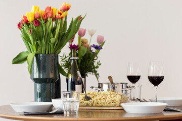 Obraz na płótnie Canvas Home-made Swabian Spaetzle (German Spätzle) in transparent glass serving dish, on a set table with tulips and other flowers, and red wine served on tall wine glasses. Small family meal for Sunday