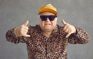 Cool retired gangsta granddad flexing to rap music. Portrait of funny weird active rich senior man in baseball cap, trendy eyeglasses and leopard patterned party shirt posing and having fun in studio - obrazy, fototapety, plakaty