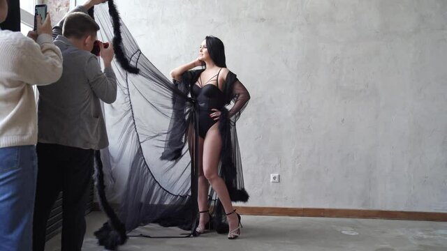sexy woman in bodysuit and negligee stands at gray wall. backstage photo shoot