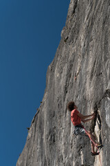 Young man in a red t-shirt and blue shorts climbs on a gray rock with a blue sky in the background.