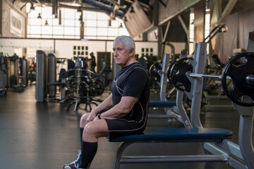 Fototapeta na wymiar an elderly man in sportswear at a workout in a fitness club sits on a bench press getting ready for an exercise