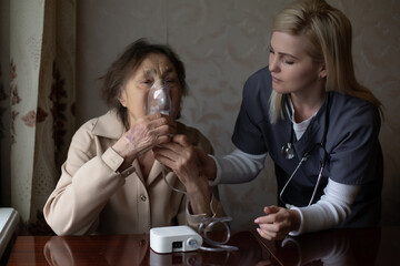 Young high-skilled pleasant woman doctor putting on nebulizer mask on face of her elderly female...