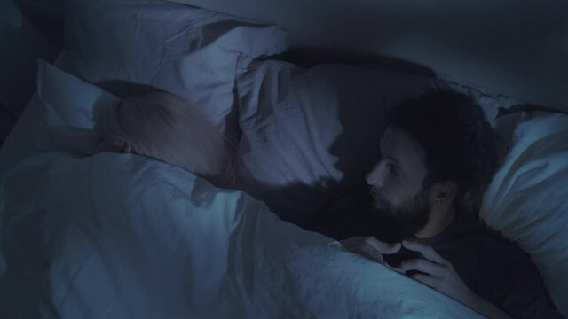Internet betrayal. Night virtual flirt. Marital disloyalty. Sneaky unfaithful husband hiding mobile phone watching video content for adults in bed with sleeping wife in dark bedroom.