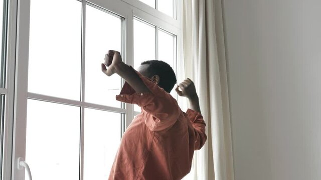 African-American black man wakes up and stretches pleasantly standing at a large bright window with curtains in the early morning before breakfast and going to work, exercises after waking up