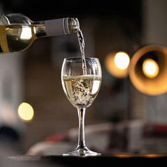White wine pours from bottle into wine glass. Alcoholic beverage with air bubbles on table against of backlit blurred background. Side view.