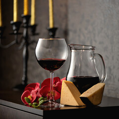 Glass jug and wine goblet with red wine, red orchid flowers and cheese on vintage candlestick background. Alcoholic drink.