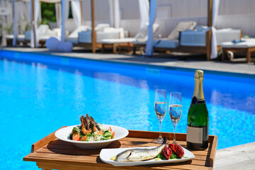 Champagne with two glasses seafood and cesar salad served on poolside on wooden tray. Luxury lifestyle concept.