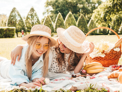 Two young beautiful smiling hipster female in summer sundress and hats.Carefree women making picnic outside.Positive models sitting on plaid on grass,drinking champagne, eating fruits and cheese