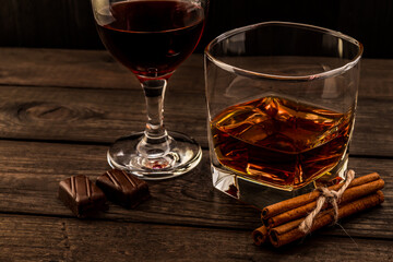 Glass of brandy and red wine with chocolate and cinnamon sticks tied with jute rope on an old wooden table. Angle view, focus on the cinnamon sticks