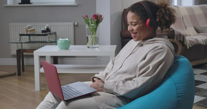 Beautiful smiling African American woman in headphones sitting on bag chair waving talking. Side view of positive confident young manager messaging online from home office. Cinema 4k ProRes HQ