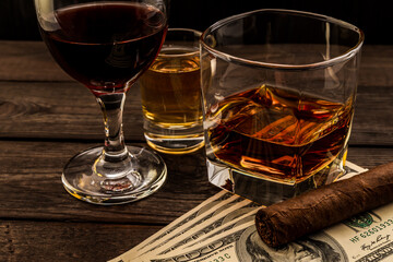 Three glasses with brandy, tequila and red wine with money and cuban cigar on an old wooden table. Angle view, focus on the cuban cigar