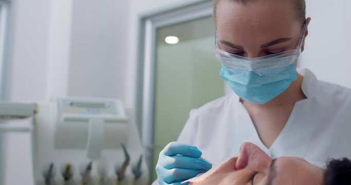 Close-up a male patient is laying in the dental chair with an optragate while a female dentist is smearing the patient&rsquo;s teeth by using a plastic stick with liquid, dental holder is in the background
