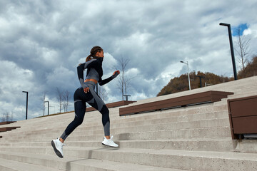 Fototapeta na wymiar Young woman runner athlete in dark skinny suit running down the city stairs, jogging and jogging at city training workout