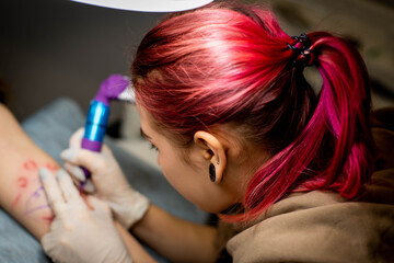 Fototapeta premium Charming girl with pink hair and tattoos stuffs a tattoo on a woman's hand in a tattoo parlor, creative small business and tattoo master. Hipster woman in gloves draws a tattoo