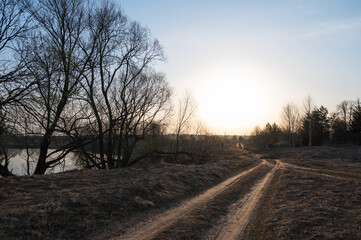 Fototapeta na wymiar Rural dirt road in a field in early spring in the rays of the rising sun against the background of bare trees and the river. Spring landscape