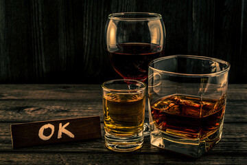 Three glasses with brandy, tequila and red wine with the wooden plank on it is an inscription "OK" on an old wooden table. Angle view focus on the inscription