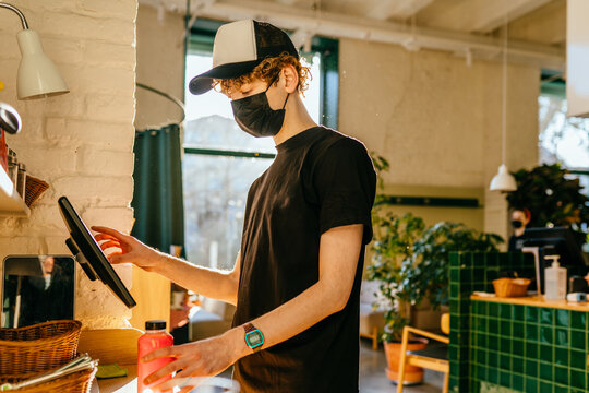 Handsome Cashier In Protective Mask Using Digital Device For Payment. A Curly Male Waiter In Uniform Calculating Someone In Coffee Shop. The Concept Of The Restaurant Business And Service Maintenance.