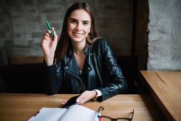 Portrait of happy Caucaisan blogger with modern cellphone device posing at cafeteria table with education textbook, cheerful female student 20s smiling at camera during leisure in coffee shop