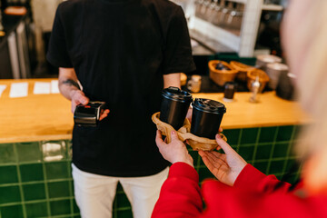 Unrecognizable man in black t-shirt standing at the counter with freshly smoothie or lemonade bottle and using terminal for contactless payment while customer holding mobile phone.