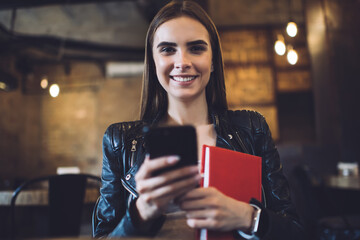 Half length portrait of cheerful hipster girl with literature book and smartphone in hands smiling at camera, funny Caucaisasn female student holding education textbook and mobile phone posing