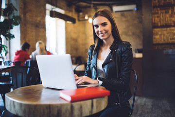 Portrait of cheerful millennial copywriter smiling at camera while doing remote work, happy Caucasian woman with laptop computer sitting at cafeteria table and posing while creating content