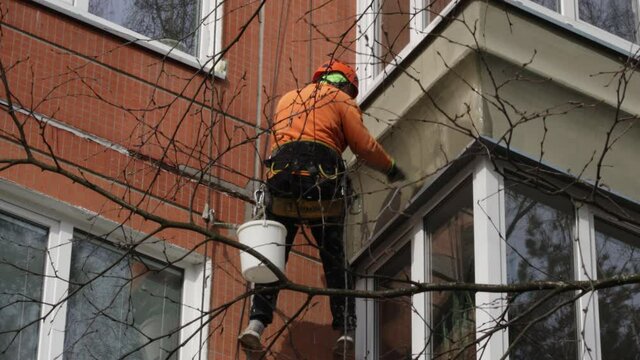 Dirty industrial climber in orange clothes working near buildings outside. Worker hanging on ropes and painting the wall with sealant. House renovation
