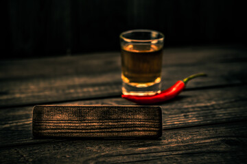 Glass of tequila with cayenne pepper and the empty wooden plank on an old wooden table. Angle view, focus on the wooden plank