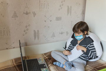 Distance learning for children during the coronavirus epidemic. The boy sits on the bed and receives a school assignment using the Internet and a laptop.