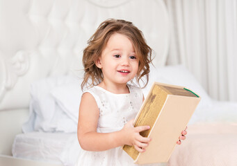 Cute little girl is holding a book in her hands. She is wearing a simple white dress. Bright room. European race.