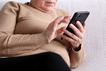 Elderly woman with smartphone sitting on sofa at home, mobile phone in female hands close up. Concept of online communication in retirement, sms, social media