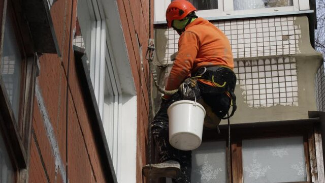 Dirty industrial climber in orange clothes working near buildings outside. Worker hanging on ropes and painting the wall with sealant. House renovation