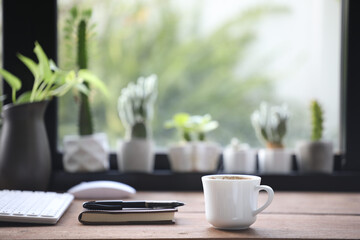 White coffee cup and notebooks and keyboard and mouse on wooden table with cactus pot
