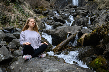 Girl sits on a large stone and meditates near a waterfall next to a mountain river.