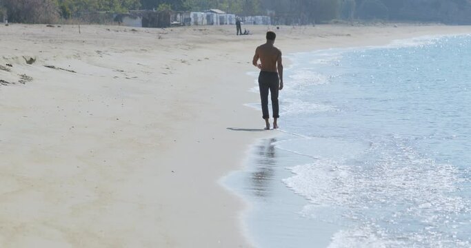 The handsome man with a perfect athletic body in gray trousers having fun on a deserted beach in the morning, He slowly runs on a beach, showing off his body and posing