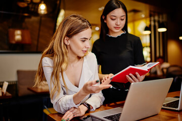Focused diverse women checking notebook and laptop in cafe