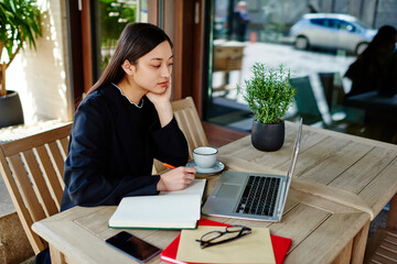 Pensive Asian student browsing netbook and writing notes in notebook on terrace