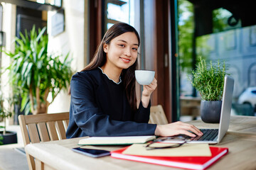 Young ethnic businesswoman drinking coffee and using laptop in cafeteria