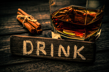 Glass of brandy with cinnamon sticks tied with jute rope and the wooden plank on it is an inscription "DRINK" on an old wooden table. Close up view, focus on the inscription