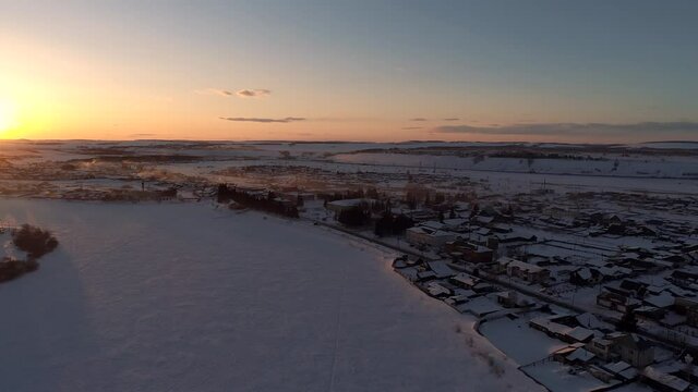Aerial over the frozen winding river surrounded with forest and with a little village on a side, Siberia, at sunset