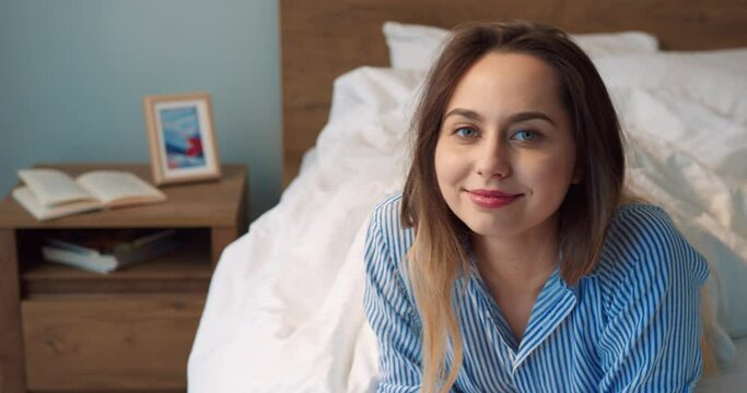 Portrait view of the calm blonde woman sitting at the bed and looking at the camera with pleasure smile while demonstrating good emotions at home at the weekend. Morning concept